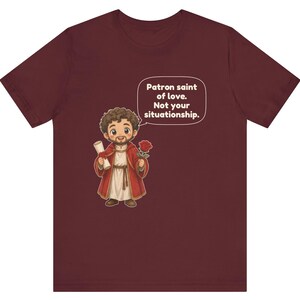 Puede incluir: Una camiseta color granate con una ilustraci&oacute;n de dibujos animados de una figura barbuda con t&uacute;nica roja que sostiene un pergamino y una rosa roja. Un globo de texto sobre la figura contiene el texto: Patron saint of love. Not your situationship.