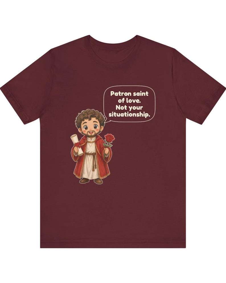 Puede incluir: Una camiseta color granate con una ilustraci&oacute;n de dibujos animados de una figura barbuda con t&uacute;nica roja que sostiene un pergamino y una rosa roja. Un globo de texto sobre la figura contiene el texto: Patron saint of love. Not your situationship.