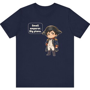 May include: A navy blue t-shirt featuring a cartoon illustration of a small figure dressed in a historical military uniform with a bicorne hat. A speech bubble next to the character contains the text: Small emperor. Big plans.