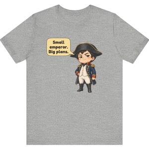 May include: A heather gray t-shirt featuring a cartoon illustration of a character dressed in a historical military uniform with a bicorne hat. A speech bubble next to the character contains the text: Small emperor. Big plans.