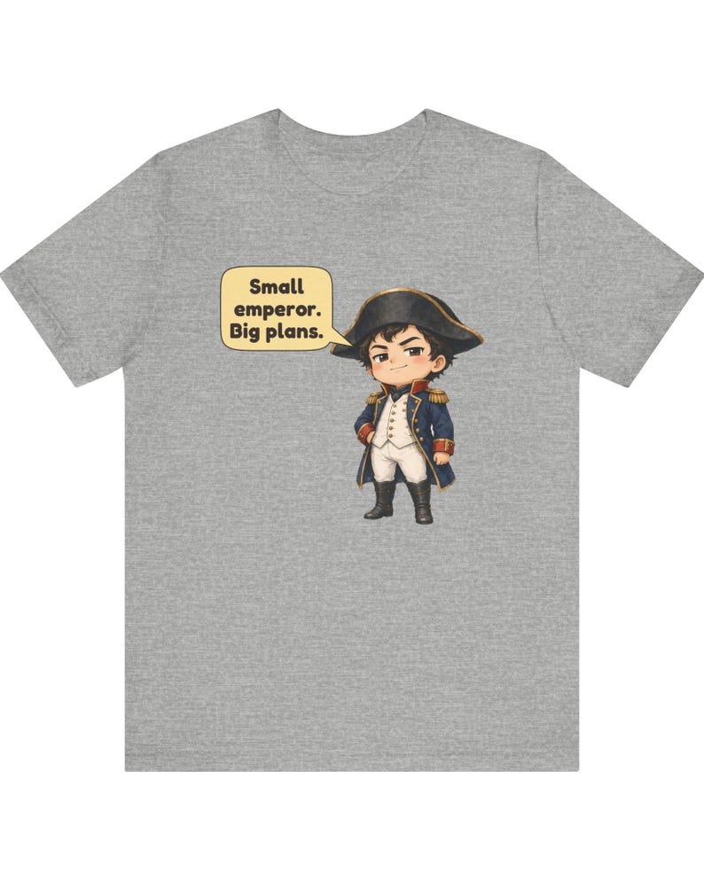May include: A heather gray t-shirt featuring a cartoon illustration of a character dressed in a historical military uniform with a bicorne hat. A speech bubble next to the character contains the text: Small emperor. Big plans.