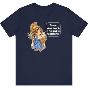 Puede incluir: Una camiseta azul marino con un gr&aacute;fico de un personaje con casco dorado y t&uacute;nica azul, sosteniendo una rama de olivo y un portapapeles con un b&uacute;ho en el hombro. Un bocadillo de di&aacute;logo contiene el texto: Show your work. The owl is watching.