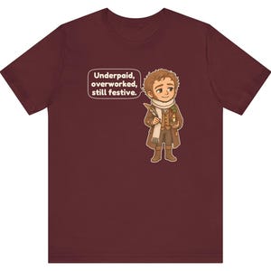 May include: A maroon short-sleeve t-shirt featuring a cartoon illustration of a person in a brown coat and white scarf holding a quill and candle. A speech bubble above the figure contains the text: Underpaid, overworked, still festive.