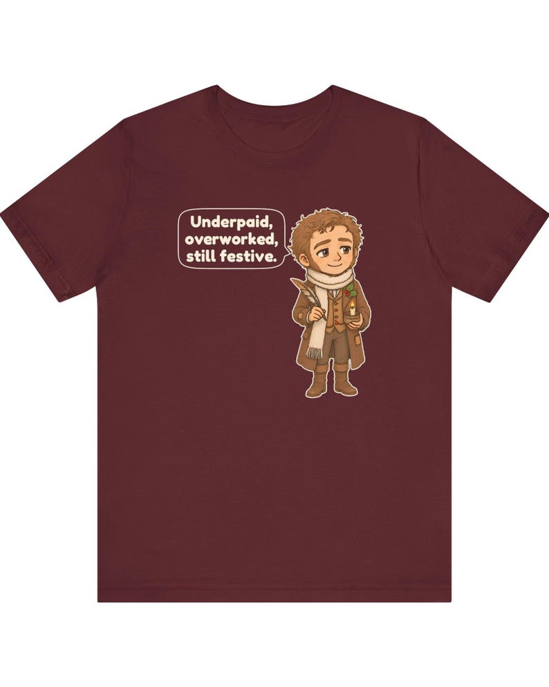 May include: A maroon short-sleeve t-shirt featuring a cartoon illustration of a person in a brown coat and white scarf holding a quill and candle. A speech bubble above the figure contains the text: Underpaid, overworked, still festive.