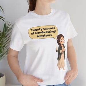 Op de afbeelding: Een wit T-shirt met korte mouwen voorzien van een cartoonillustratie van een persoon die een kaars vasthoudt en een tekstballon met de tekst: Twenty seconds of handwashing? Amateurs. De afbeelding staat op de borst van het shirt.