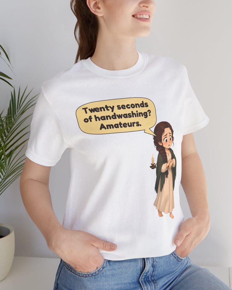 Op de afbeelding: Een wit T-shirt met korte mouwen voorzien van een cartoonillustratie van een persoon die een kaars vasthoudt en een tekstballon met de tekst: Twenty seconds of handwashing? Amateurs. De afbeelding staat op de borst van het shirt.