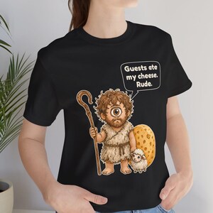 May include: A black t-shirt featuring a graphic of a cyclops holding a wooden staff next to a small white sheep and a large piece of Swiss cheese. A speech bubble above the cyclops contains the text: Guests ate my cheese. Rude.