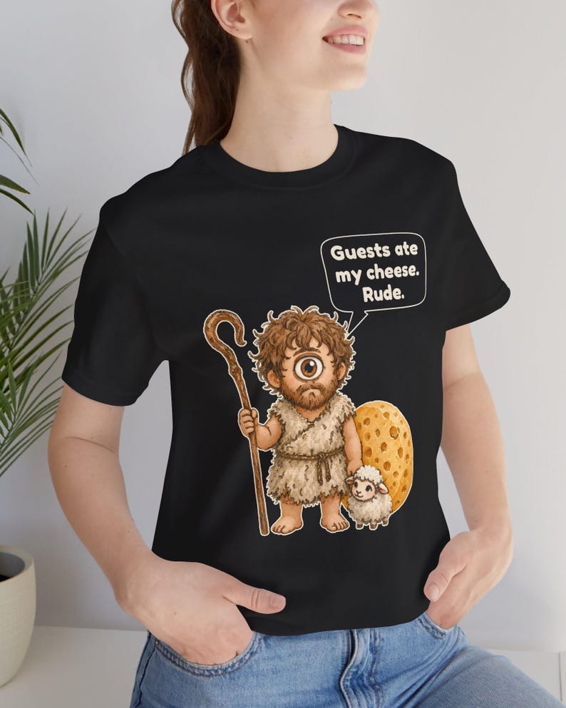 May include: A black t-shirt featuring a graphic of a cyclops holding a wooden staff next to a small white sheep and a large piece of Swiss cheese. A speech bubble above the cyclops contains the text: Guests ate my cheese. Rude.