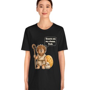 May include: A black t-shirt featuring a graphic of a one-eyed cyclops holding a wooden staff and standing next to a small sheep and a large wedge of Swiss cheese. A speech bubble above the cyclops contains the text: Guests ate my cheese. Rude.