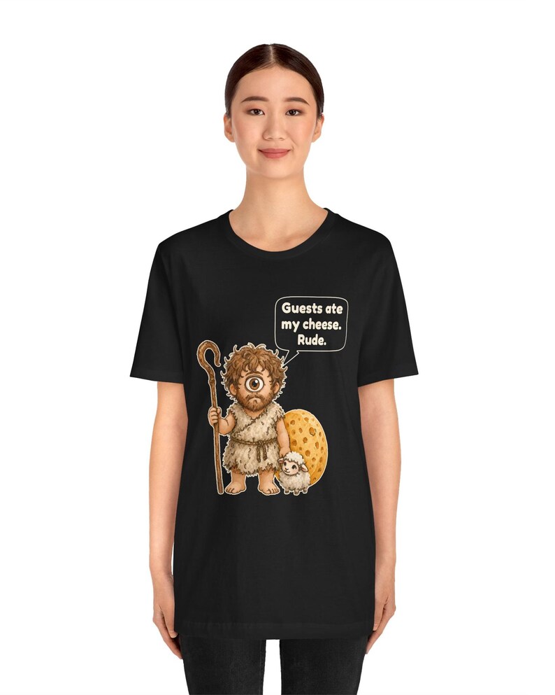 May include: A black t-shirt featuring a graphic of a one-eyed cyclops holding a wooden staff and standing next to a small sheep and a large wedge of Swiss cheese. A speech bubble above the cyclops contains the text: Guests ate my cheese. Rude.