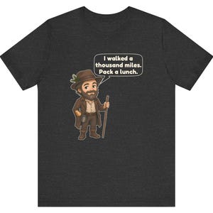May include: A dark charcoal gray t-shirt featuring a cartoon illustration of a bearded hiker wearing a brown hat with a pine sprig, a brown coat, and boots, holding a wooden walking stick, with a speech bubble saying I walked a thousand miles. Pack a lunch.