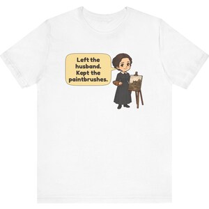 May include: A white t-shirt featuring a cartoon illustration of a person in a black dress standing next to an easel with a painting. A speech bubble contains the text: Left the husband. Kept the paintbrushes.