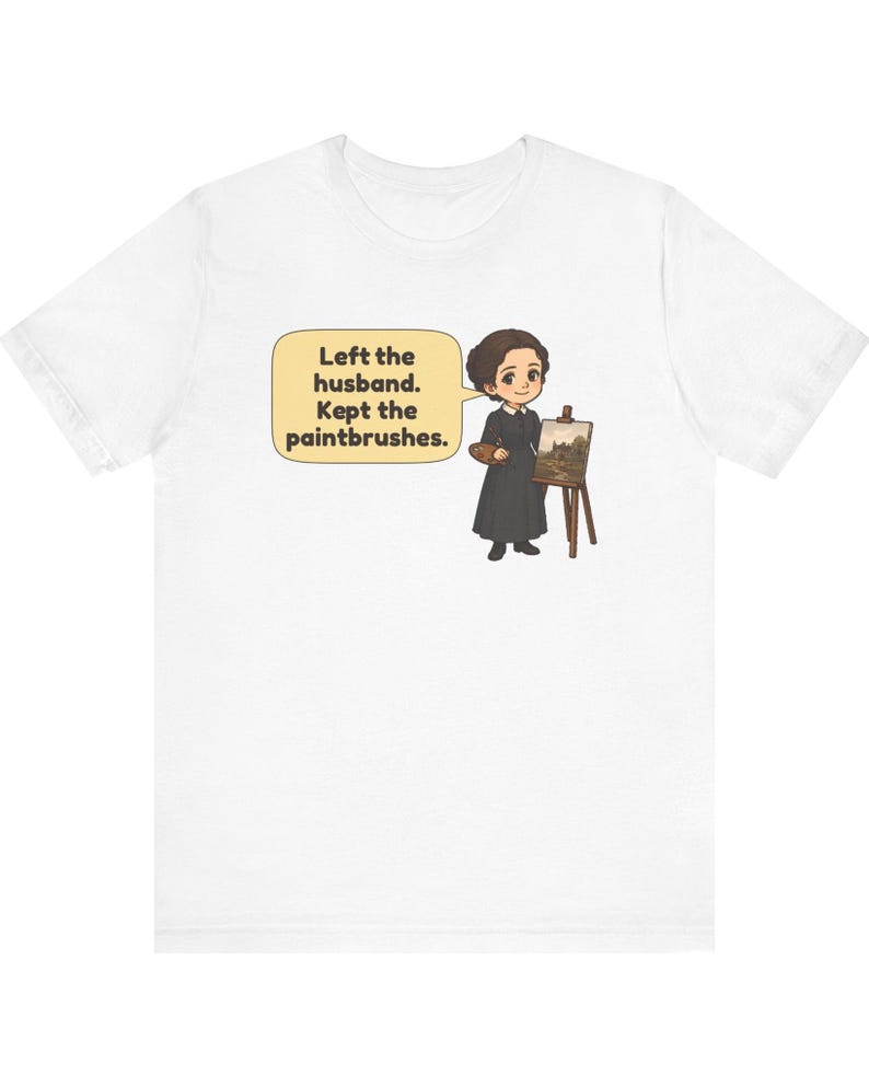 May include: A white t-shirt featuring a cartoon illustration of a person in a black dress standing next to an easel with a painting. A speech bubble contains the text: Left the husband. Kept the paintbrushes.