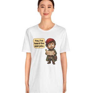May include: A white t-shirt featuring a cartoon illustration of a bearded character wearing a red cap and green coat, holding a map. A speech bubble next to the character contains the text, "Yes, I've heard the pool joke." The design is centred on the chest.