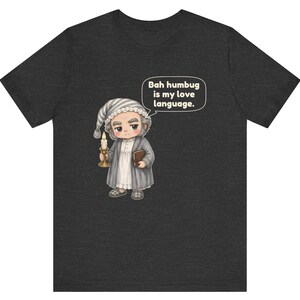 Puede incluir: Una camiseta gris jaspeado oscuro con una ilustraci&oacute;n de dibujos animados de un personaje con gorro de dormir y bata que sostiene una vela y un libro. Un globo de texto sobre el personaje contiene el texto, Bah humbug is my love language.
