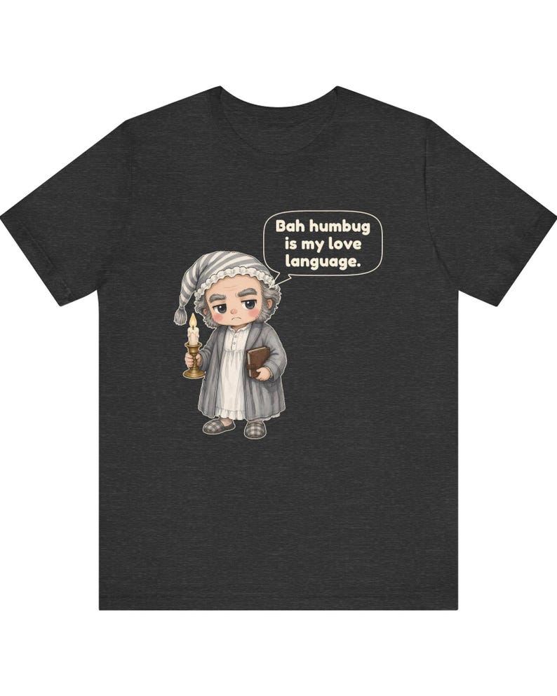 Puede incluir: Una camiseta gris jaspeado oscuro con una ilustraci&oacute;n de dibujos animados de un personaje con gorro de dormir y bata que sostiene una vela y un libro. Un globo de texto sobre el personaje contiene el texto, Bah humbug is my love language.