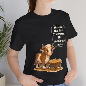 以下が含まれることがあります： 黒の半袖Tシャツ。干し草が入った木製の飼い葉桶の横に立つ、茶色と白の子牛のグラフィックが描かれています。子牛の上の吹き出しには「Hosted the first Christmas. No thank-you note.」というテキストが書かれています。