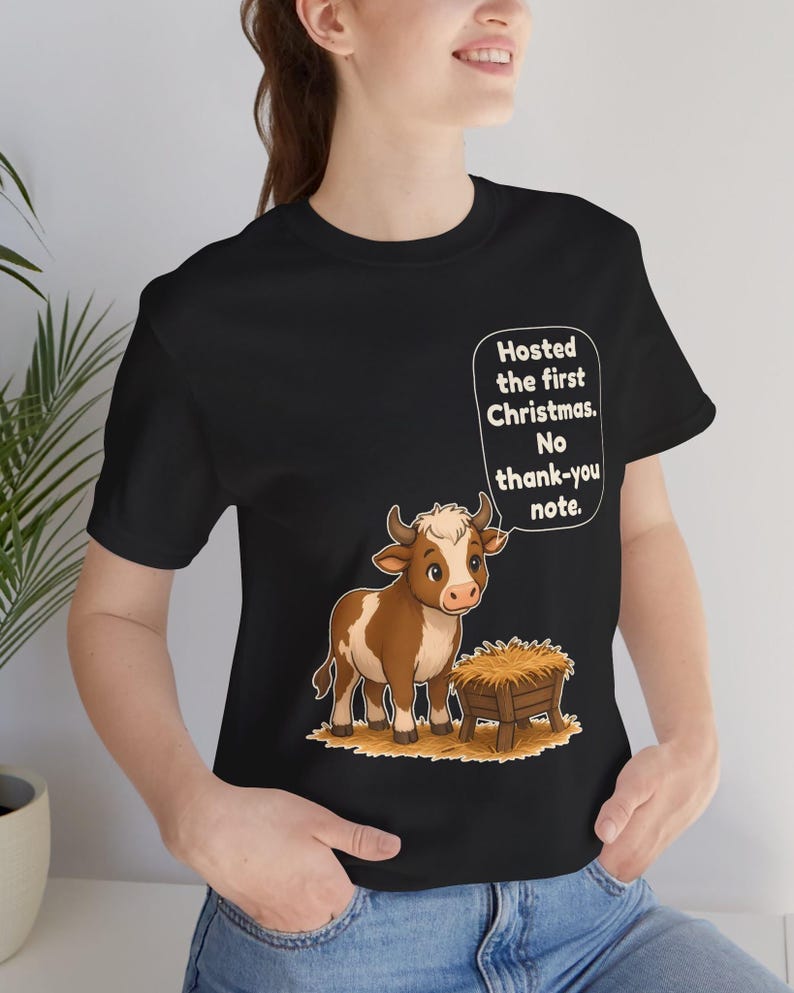以下が含まれることがあります： 黒の半袖Tシャツ。干し草が入った木製の飼い葉桶の横に立つ、茶色と白の子牛のグラフィックが描かれています。子牛の上の吹き出しには「Hosted the first Christmas. No thank-you note.」というテキストが書かれています。