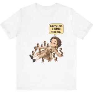 Op de afbeelding: Een wit t-shirt met een afbeelding van een groot persoon die ligt, omringd door vele kleine mensen die touwen en ladders gebruiken om hen vast te binden. Een gele tekstballon bevat de tekst, Sorry, I'm a little tied up.