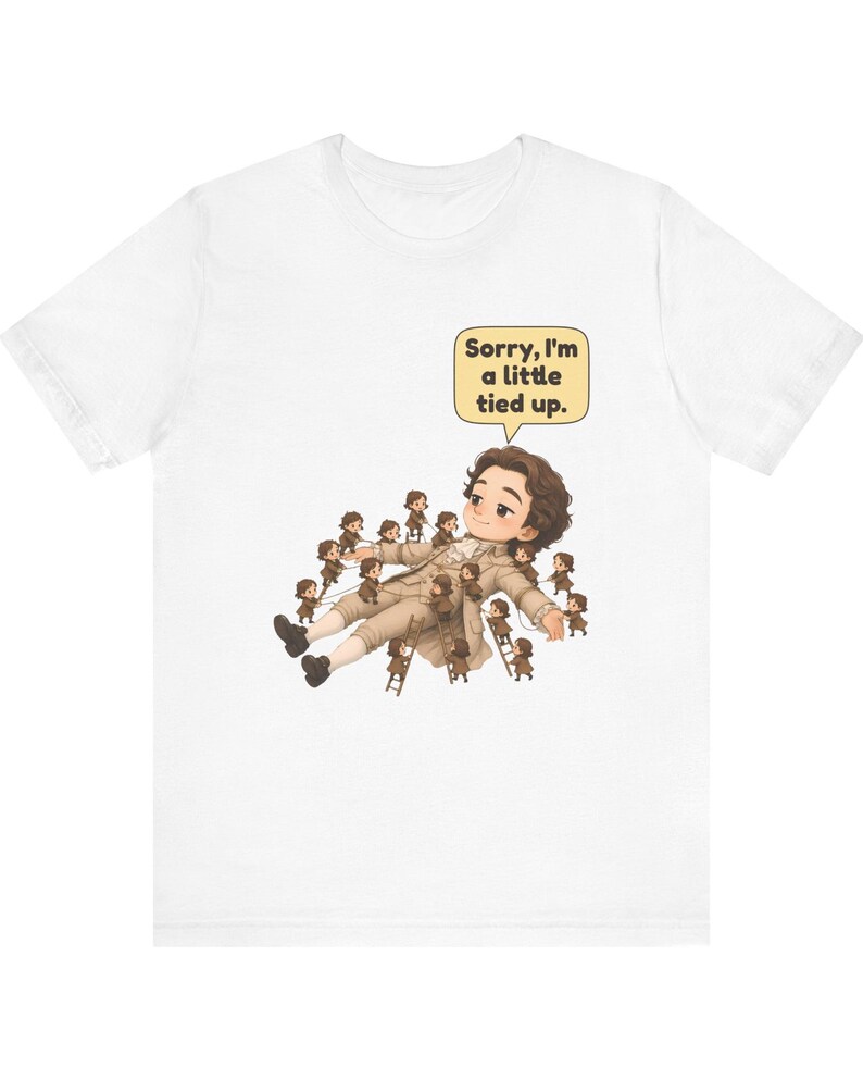 Op de afbeelding: Een wit t-shirt met een afbeelding van een groot persoon die ligt, omringd door vele kleine mensen die touwen en ladders gebruiken om hen vast te binden. Een gele tekstballon bevat de tekst, Sorry, I'm a little tied up.