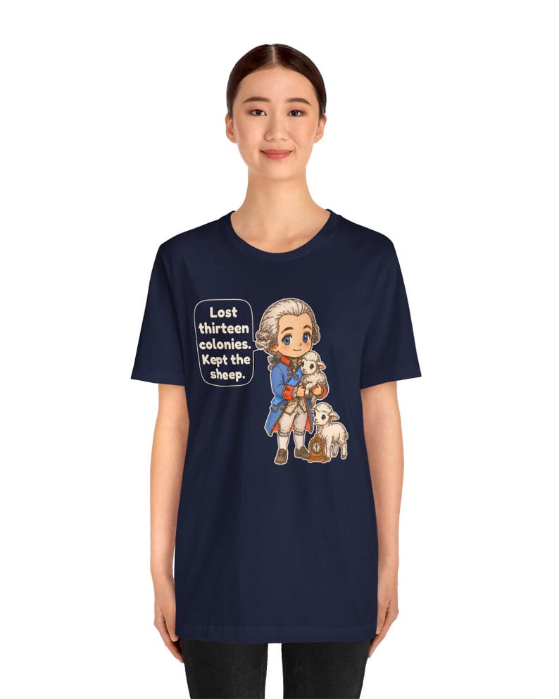 Peut inclure: Un t-shirt bleu marine &agrave; manches courtes avec une illustration de style dessin anim&eacute; repr&eacute;sentant un personnage historique en manteau bleu tenant un agneau blanc, avec un autre agneau &agrave; c&ocirc;t&eacute;. Une bulle contient le texte : Lost thirteen colonies. Kept the sheep.