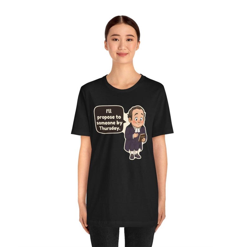 May include: A black t-shirt featuring a cartoon illustration of a man in a dark coat and white cravat holding a small book. A speech bubble next to the character contains the text, "I'll propose to someone by Thursday."