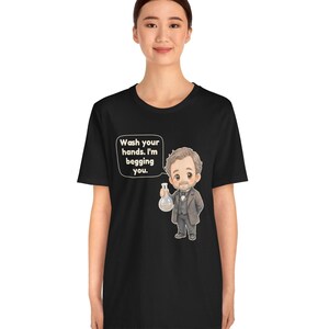 May include: A black t-shirt featuring a cartoon illustration of a man in a suit holding a glass flask. A speech bubble next to the character contains the text: Wash your hands. I'm begging you. The design is centered on the front of the shirt.