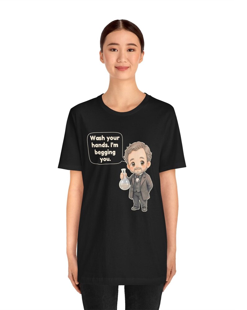 May include: A black t-shirt featuring a cartoon illustration of a man in a suit holding a glass flask. A speech bubble next to the character contains the text: Wash your hands. I'm begging you. The design is centered on the front of the shirt.