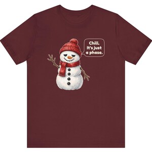 Pode incluir: Uma t-shirt de manga curta cor de vinho com o gr&aacute;fico de um boneco de neve a usar um gorro e um cachecol de malha vermelhos. O boneco de neve tem um nariz de cenoura, bra&ccedil;os de pau e um bal&atilde;o com o texto, Chill. It's just a phase.