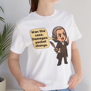 May include: A white t-shirt featuring a cartoon illustration of a historical figure in a brown suit holding a quill. A speech bubble contains the text: Won the case. Damages: pocket change. The figure has a powdered wig and is pointing upward.