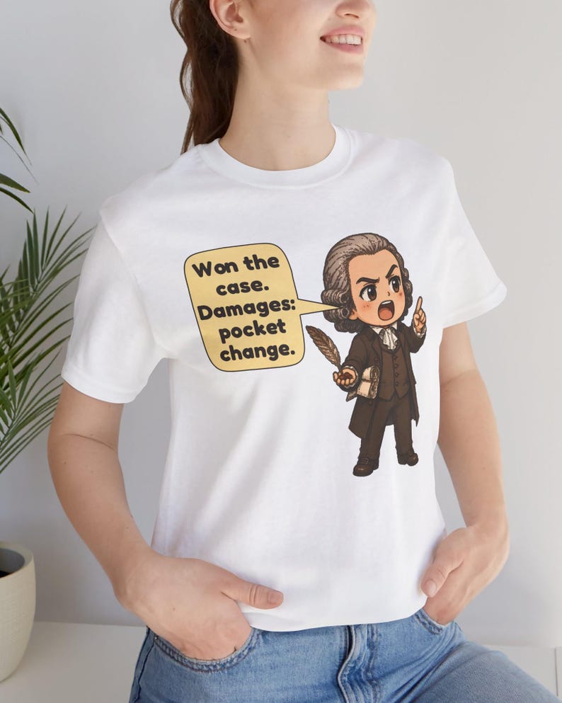 May include: A white t-shirt featuring a cartoon illustration of a historical figure in a brown suit holding a quill. A speech bubble contains the text: Won the case. Damages: pocket change. The figure has a powdered wig and is pointing upward.