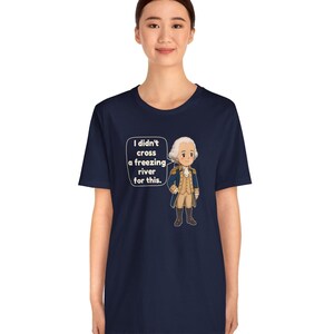 May include: A navy blue short-sleeve t-shirt featuring a cartoon illustration of a historical figure in a blue coat and white wig. A speech bubble next to the character contains the text: I didn't cross a freezing river for this.