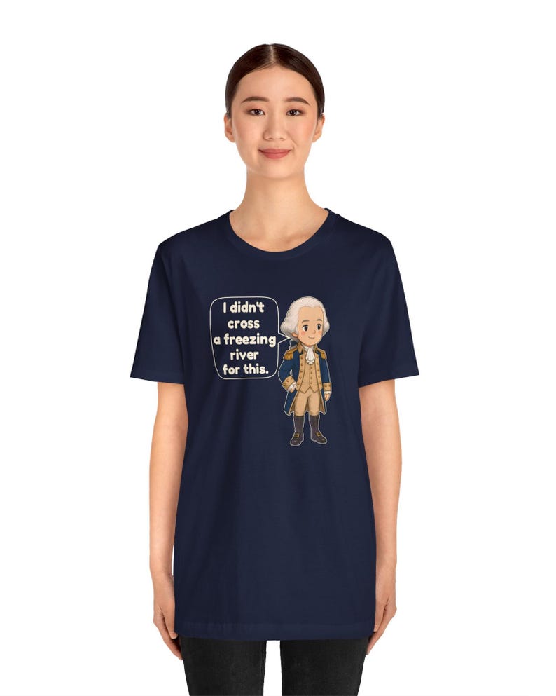 May include: A navy blue short-sleeve t-shirt featuring a cartoon illustration of a historical figure in a blue coat and white wig. A speech bubble next to the character contains the text: I didn't cross a freezing river for this.