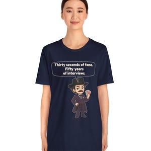 Pu&ograve; includere: Una t-shirt blu navy con un'illustrazione in stile cartoon di un uomo in abito scuro, cappello e baffi che tiene in mano un ventaglio di carte. Un fumetto sopra il personaggio contiene il testo: Thirty seconds of fame. Fifty years of interviews.