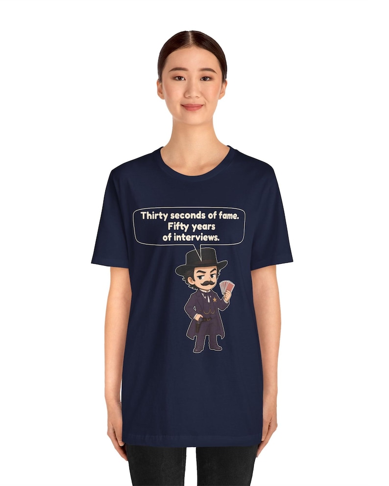 Pu&ograve; includere: Una t-shirt blu navy con un'illustrazione in stile cartoon di un uomo in abito scuro, cappello e baffi che tiene in mano un ventaglio di carte. Un fumetto sopra il personaggio contiene il testo: Thirty seconds of fame. Fifty years of interviews.