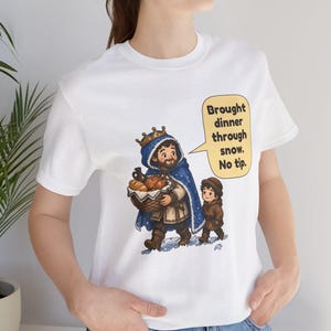 May include: A white t-shirt featuring a graphic illustration of a person wearing a blue cloak and a gold crown, carrying a basket of bread, walking with a child in the snow. A yellow speech bubble contains the text: Brought dinner through snow. No tip.