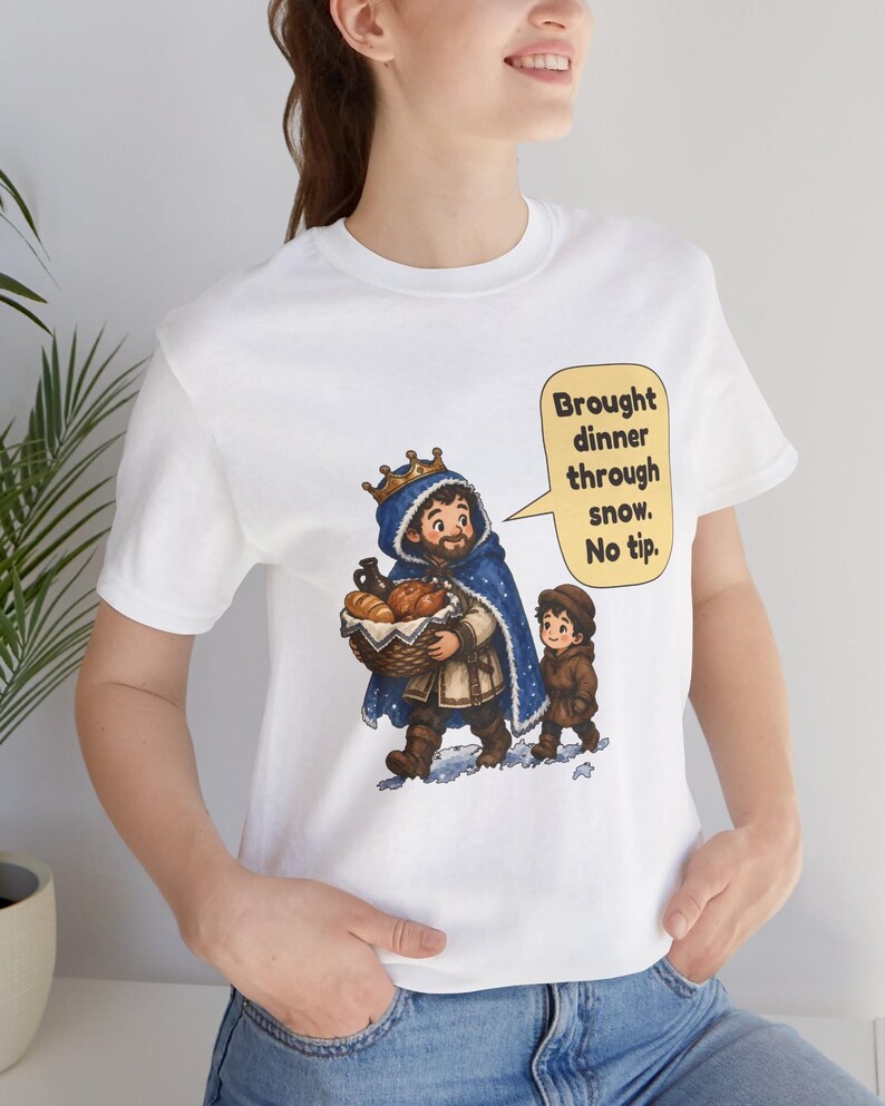 May include: A white t-shirt featuring a graphic illustration of a person wearing a blue cloak and a gold crown, carrying a basket of bread, walking with a child in the snow. A yellow speech bubble contains the text: Brought dinner through snow. No tip.