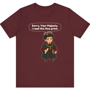 May include: A maroon short-sleeve t-shirt featuring a cartoon character wearing a black cap with a halo, a dark coat with a gold chain, and holding a scroll and book. A speech bubble above the character reads, Sorry, Your Majesty. I read the fine print.