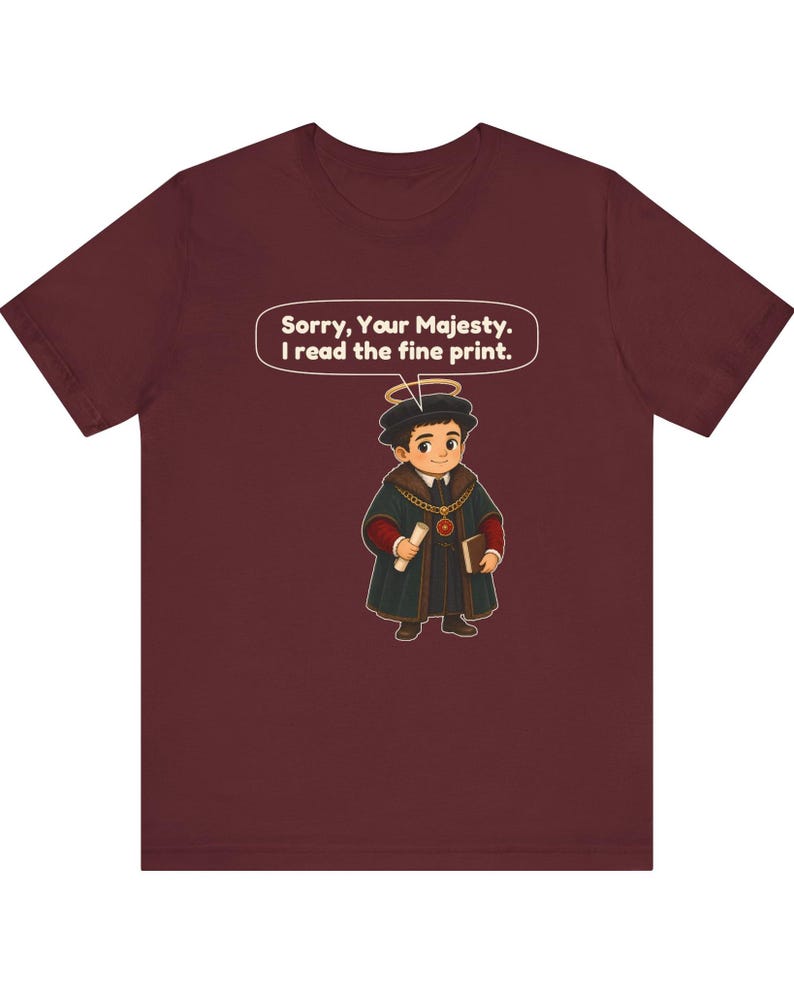 May include: A maroon short-sleeve t-shirt featuring a cartoon character wearing a black cap with a halo, a dark coat with a gold chain, and holding a scroll and book. A speech bubble above the character reads, Sorry, Your Majesty. I read the fine print.