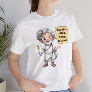 May include: A white t-shirt featuring a cartoon illustration of a bearded man with curly gray hair wearing a white toga with blue trim and brown sandals. The character holds a white towel and has a speech bubble that says, Eureka! Also, I need a towel.