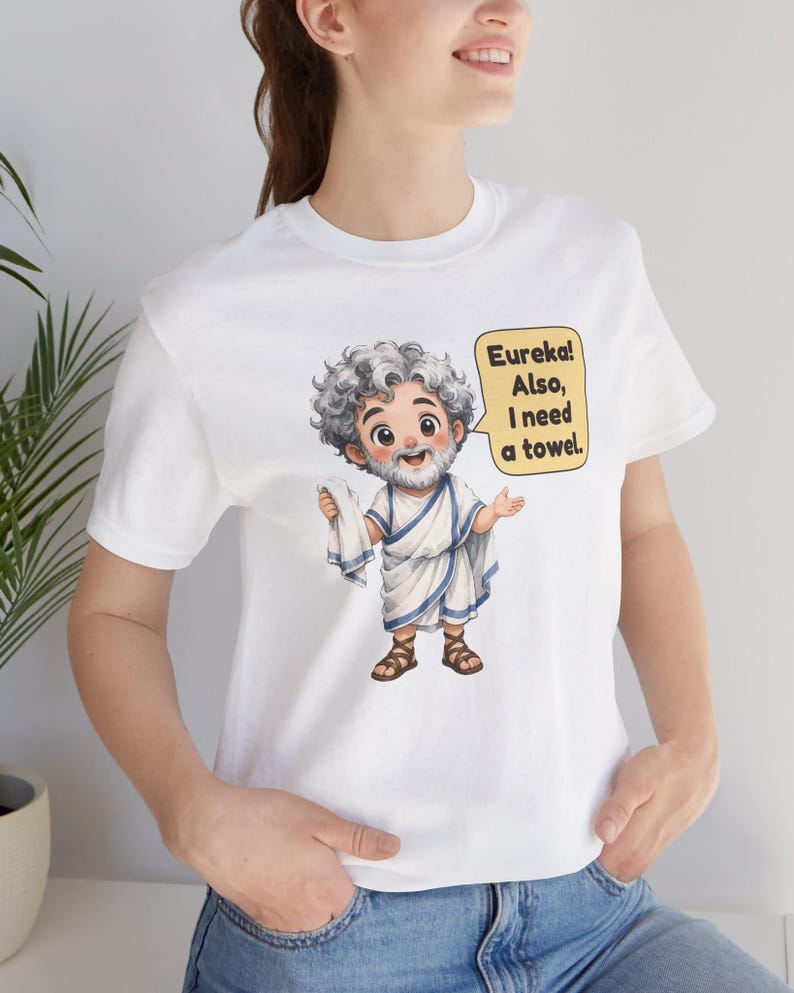 May include: A white t-shirt featuring a cartoon illustration of a bearded man with curly gray hair wearing a white toga with blue trim and brown sandals. The character holds a white towel and has a speech bubble that says, Eureka! Also, I need a towel.