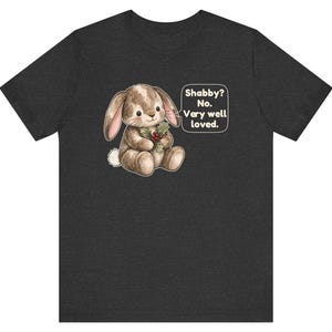 May include: A dark charcoal gray short-sleeve t-shirt featuring a brown plush rabbit illustration holding holly berries. A speech bubble next to the rabbit contains the text: Shabby? No. Very well loved. The design is centered on the chest.