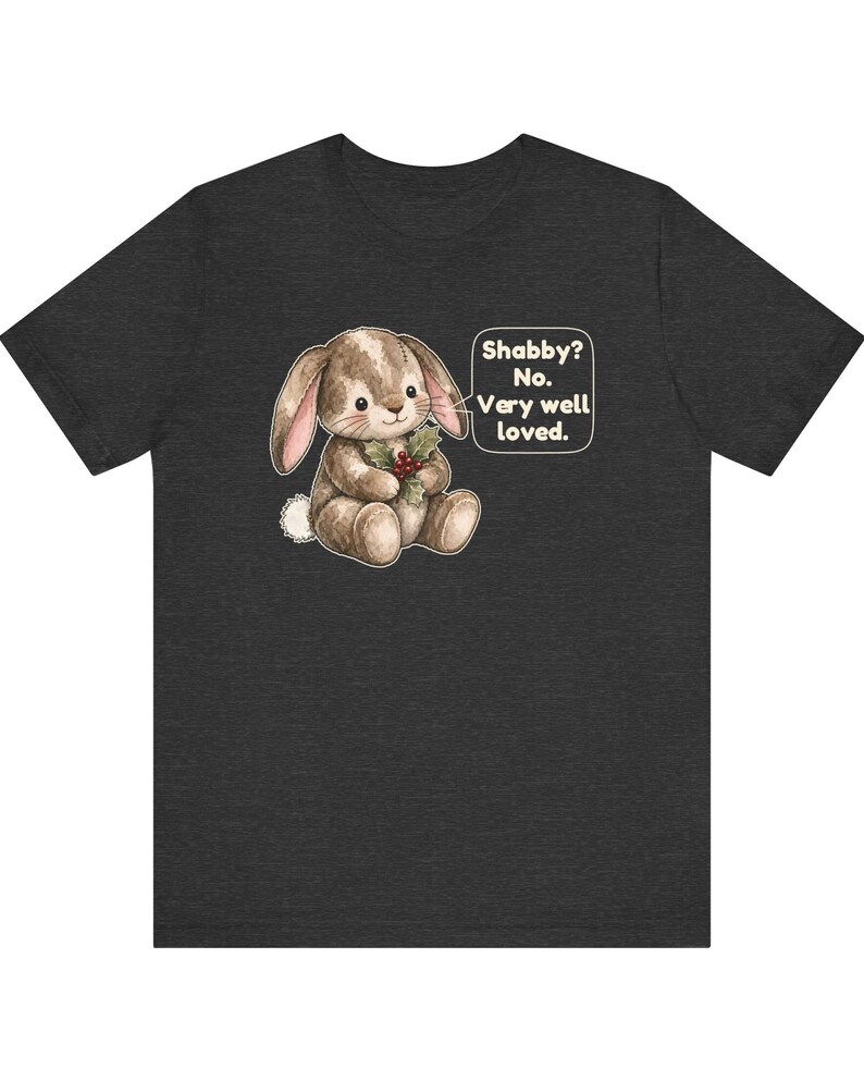 May include: A dark charcoal gray short-sleeve t-shirt featuring a brown plush rabbit illustration holding holly berries. A speech bubble next to the rabbit contains the text: Shabby? No. Very well loved. The design is centered on the chest.