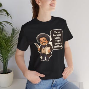 May include: A black t-shirt featuring a cartoon illustration of a man with an afro and mustache wearing a brown suit and bow tie. He holds a quill and paper. A speech bubble contains the text: Three hundred books. Then I wrote a cookbook.