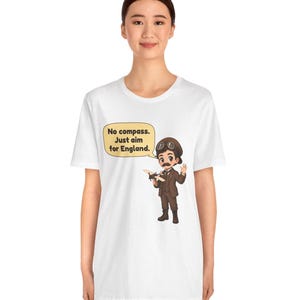 May include: A white t-shirt featuring a cartoon illustration of a pilot wearing a brown suit, cap, and goggles, holding a small airplane. A speech bubble next to the character contains the text: No compass. Just aim for England.