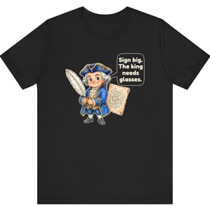 May include: A black t-shirt featuring a cartoon illustration of a historical figure in a blue coat and tricorn hat holding a white quill and a scroll. A speech bubble contains the text: Sign big. The king needs glasses.