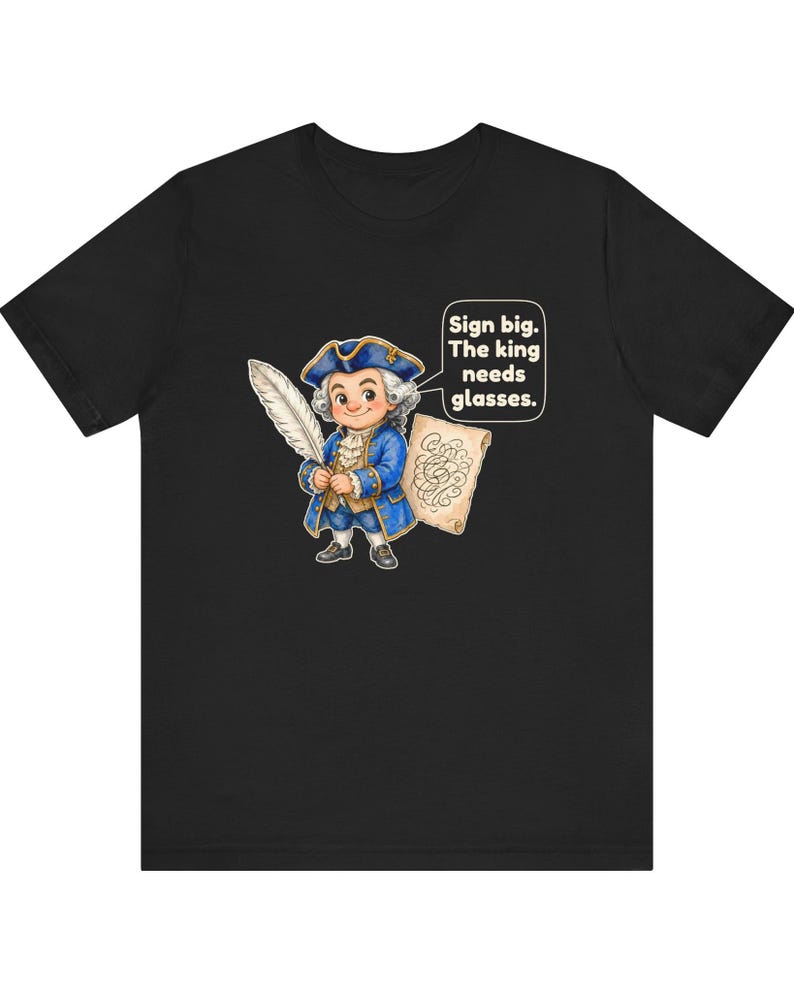 May include: A black t-shirt featuring a cartoon illustration of a historical figure in a blue coat and tricorn hat holding a white quill and a scroll. A speech bubble contains the text: Sign big. The king needs glasses.