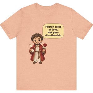 Puede incluir: Una camiseta color melocot&oacute;n con una ilustraci&oacute;n de dibujos animados de una figura barbuda con t&uacute;nica roja que sostiene un pergamino y una rosa roja. Un bocadillo sobre la figura contiene el texto: Patron saint of love. Not your situationship.