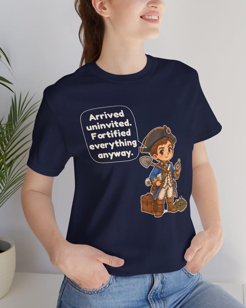 May include: A navy blue t-shirt featuring a graphic of a cartoon character in a historical military uniform, holding a shovel and compass, standing next to a wooden chest. A speech bubble contains the text: Arrived uninvited. Fortified everything anyway.