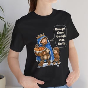 May include: A black t-shirt featuring a graphic of a bearded person wearing a blue crown and cape, carrying a basket of food, walking with a child in the snow. A speech bubble contains the text: Brought dinner through snow. No tip.
